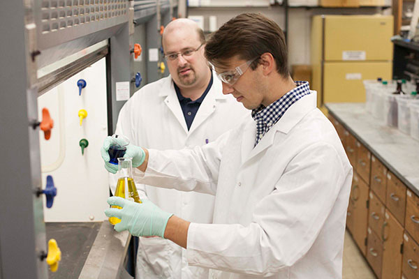 A professor observes as a student in a lab coat prepares to mix two chemicals in a flask