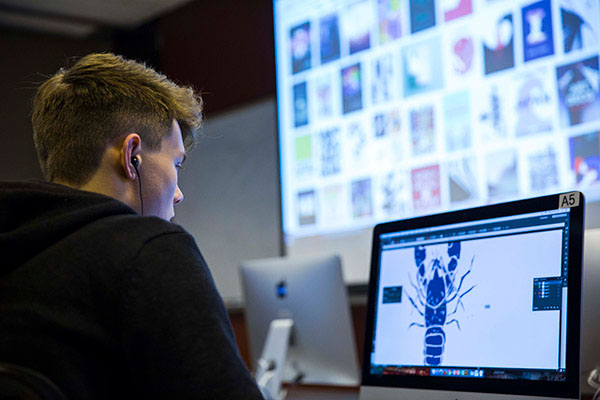 A student working on a desktop uses Adobe Illustrator to craft a logo of a lobster