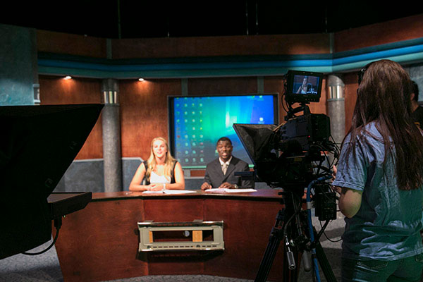 A woman stands behind a camera recording two newscasters at a desk