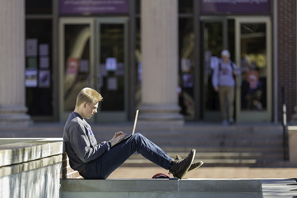 A student sits outside while completing work on his laptop
