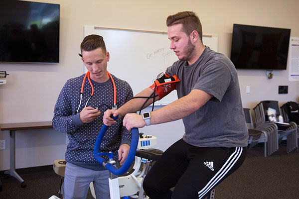 One student takes the blood pressure of another student that is on an exercise bike