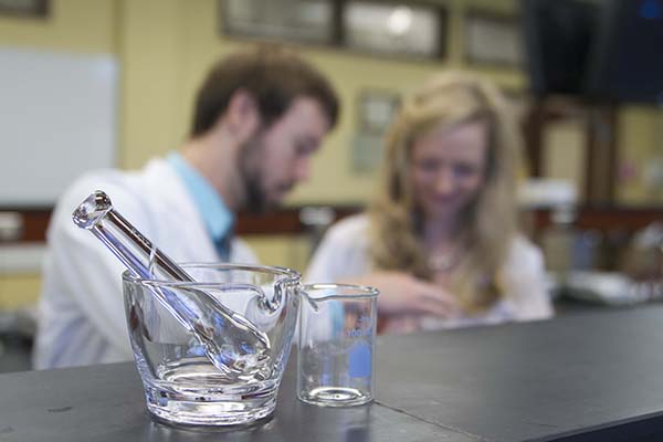 A beaker, mortar and pestle sit on a lab bench while two students conduct an experiment in the background