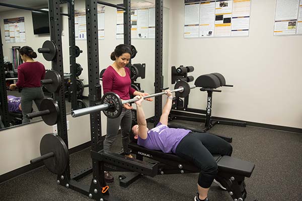 One student spots another student as she lifts a barbell