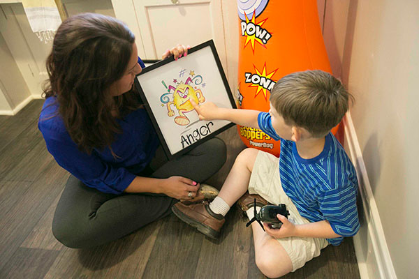 A woman sits on the floor with a young boy that points to an emotion on a chart held by the woman