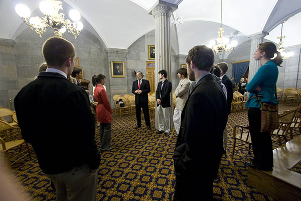 A group of students stand in a parliament building and listen to a man talk