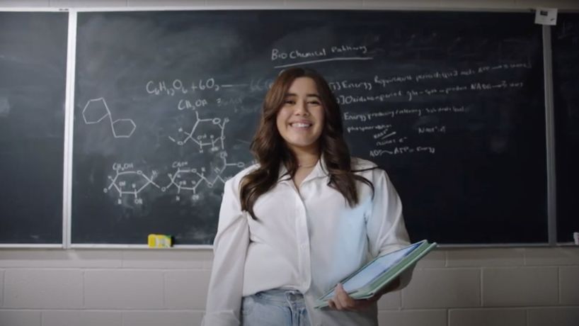 Student with a notebook in front of a chalk board smiling