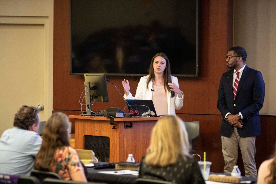 Bradley Bruce standing beside podium while another student presents her pitch.