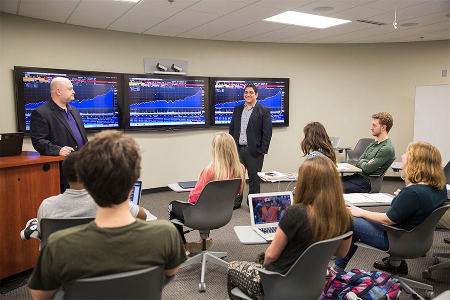 Two professors stand in front of class seated in movable desks with information on screens behind.