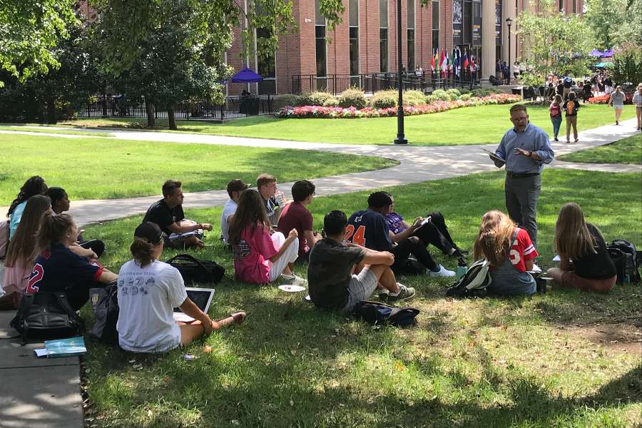 Group of students sitting under tree listening to faculty member. 