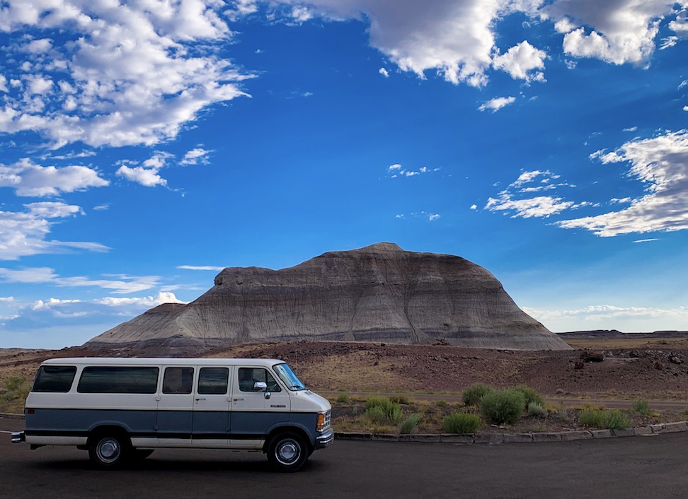 An old grey and white 15 passenger van sits in front of a large mountain and sky.
