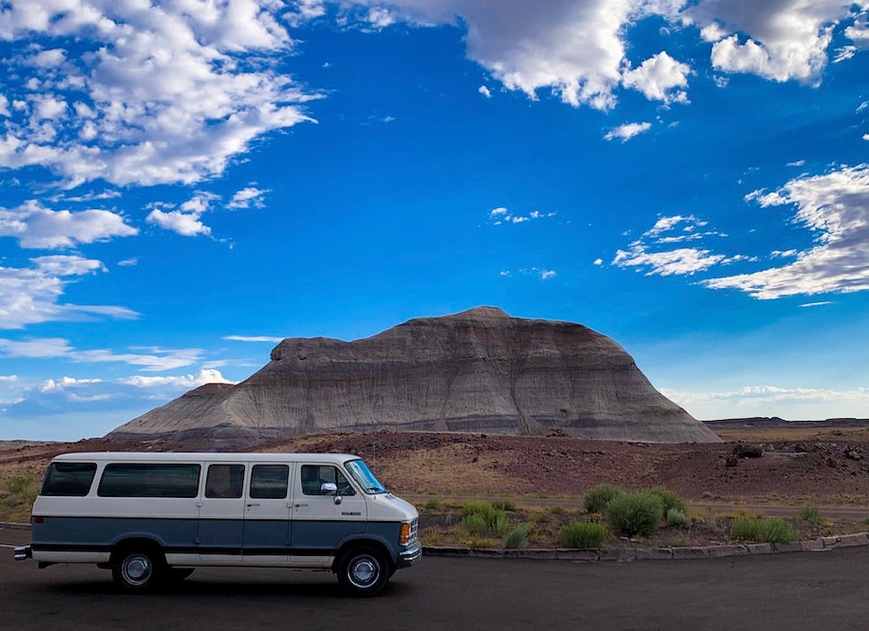 An old grey and white 15 passenger van sits in front of a large mountain and sky.