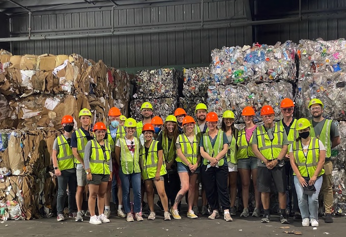 A group of students wearing high-vis vests and helmets stand in front of cubes of materials ready for recycling.