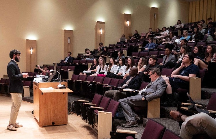 student presenting paper in lecture hall with students and professors