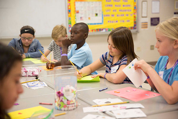 Six elementary school aged children sit at desks sketching on paper