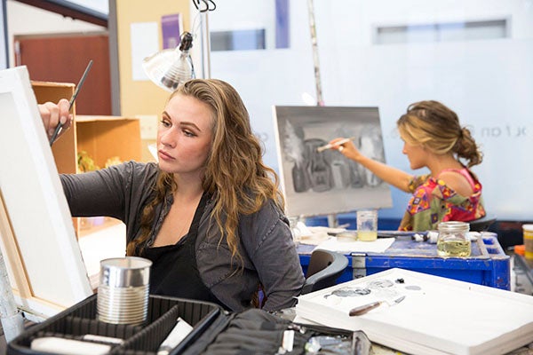 Two female students paint black and white images on canvases in a classroom