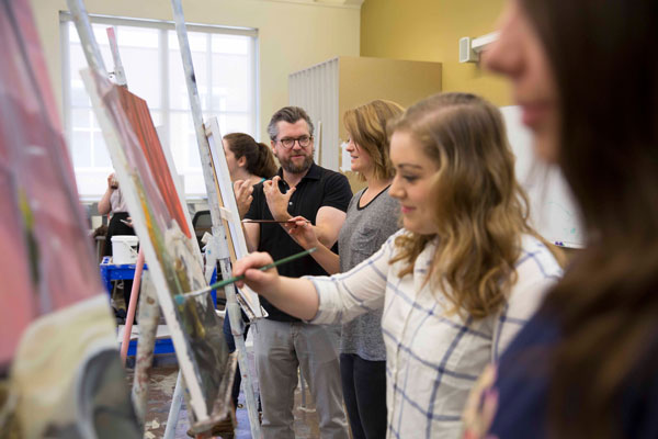 A professor stands with a student in a classroom studio and gives her advice for her painting