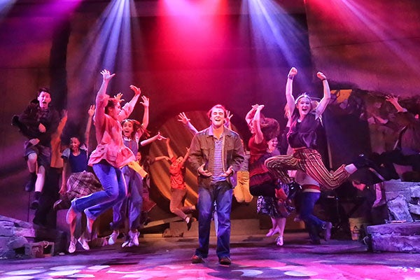 Students in a school production dance and jump around another student singing a solo