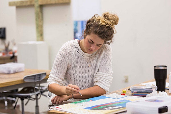 A female student sits at a work table and uses a sketch pad and colored pencils to draw an image