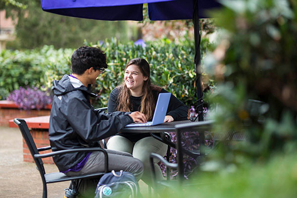 Two students sit outside laughing as one works on his laptop