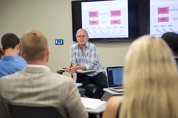 A professor sits in front of his class as he lectures