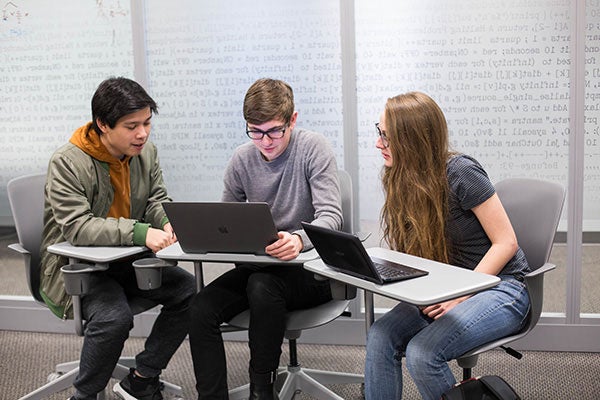 Three students study a laptop screen in a classroom