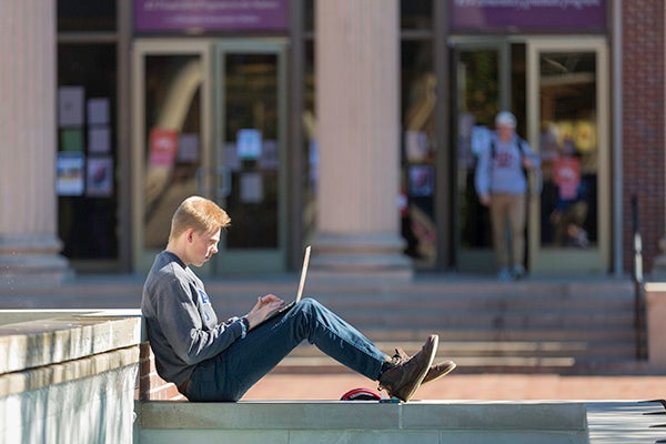 A student sits outside while completing work on his laptop