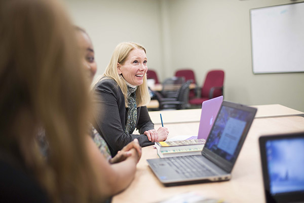 A professor sits at a table with students smiling as she takes notes on a presentation