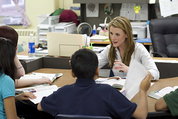 A teacher instructs three students sitting across from her using a textbook