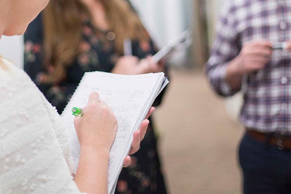 A woman takes notes in a spiral notebook as she stands across from two others