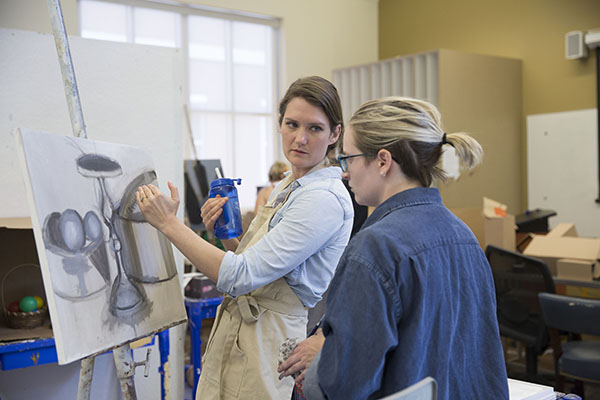 A professor stands at a canvas with a student and gestures to one of the shapes the student has drawn