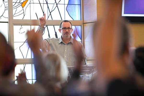 A professor points his finger upward as the the students in the classroom raise their hands.