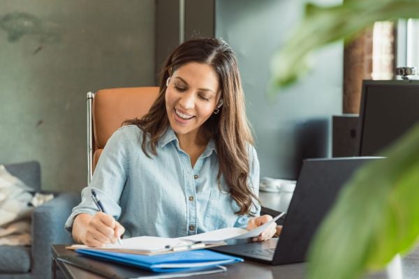 A woman writes on her notebook while sitting next to her laptop