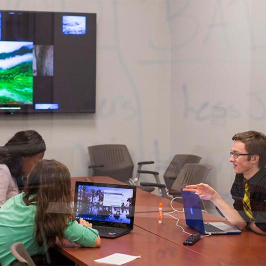 Three students gathered around a table working on a computer as seen through a glass wall with writing.